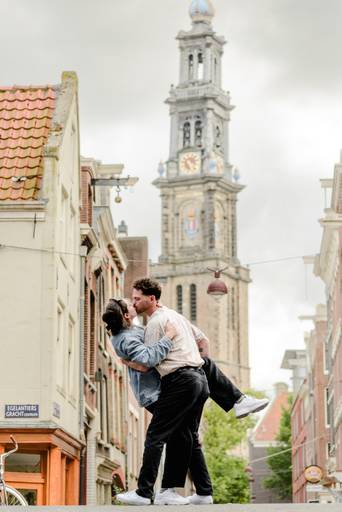 Couple stands on an Amsterdam bridge, smiling and talking right after their surprise engagement, with boats and canal houses behind them.