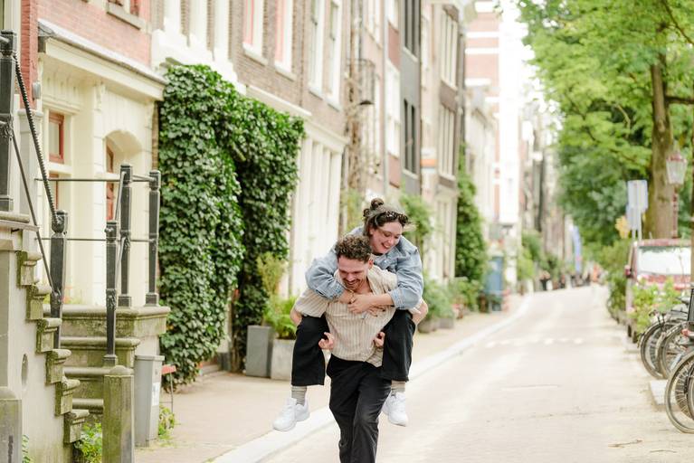 Playful engagement moment in Amsterdam as one partner gives a piggyback ride along a quiet, charming street lined with classic Dutch homes.