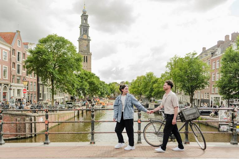 Couple holding hands and walking across an Amsterdam canal bridge, with Westerkerk in the background during their engagement photoshoot.