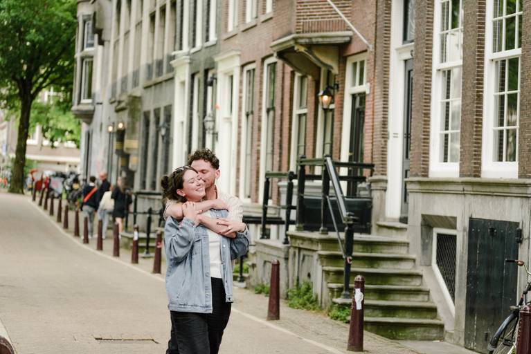 Engaged couple hugging and smiling while walking through a peaceful Amsterdam street lined with historic houses.