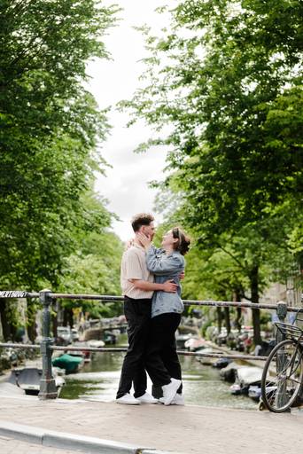 Engaged couple sharing a kiss on an Amsterdam bridge surrounded by trees and historic canals after a surprise proposal.
