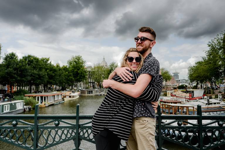 Couple hugging on a bridge with Amsterdam houseboats and canal views during their engagement session.