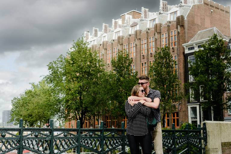 Couple hugging on a bridge in Amsterdam with historic buildings and trees in the background during their proposal session.