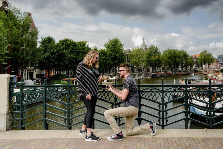 Man proposing on a canal bridge in Amsterdam while the woman smiles in surprise during their engagement shoot.