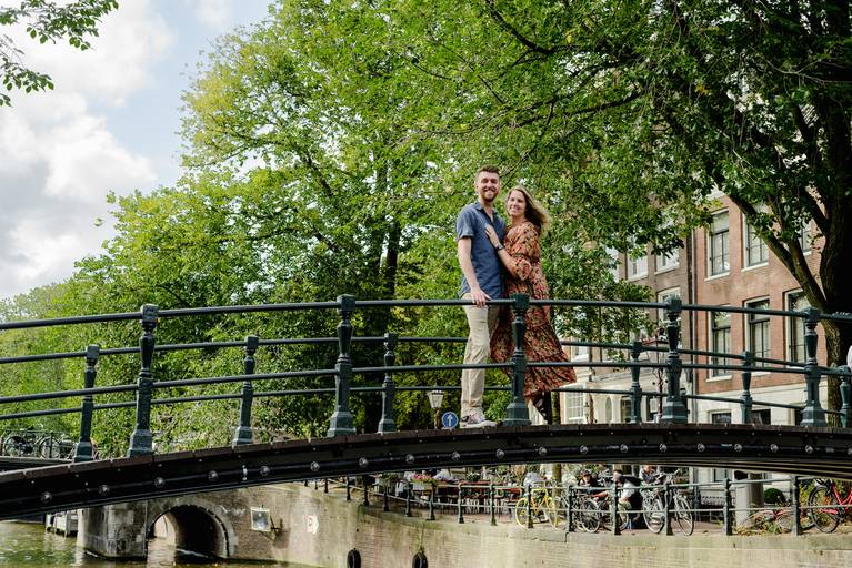 Couple standing together on a bridge in Amsterdam, surrounded by tall trees and canal views during a surprise proposal photo session.