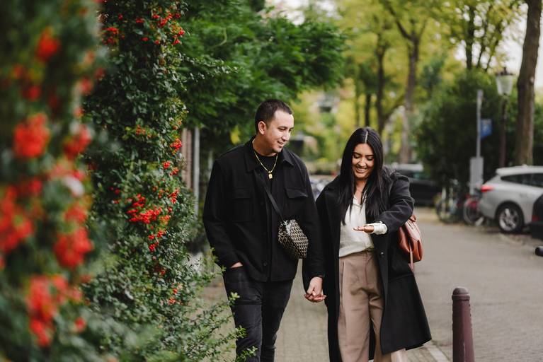 Newly engaged couple smiles at each other on an Amsterdam canal bridge with trees and boats in the background after a surprise proposal.