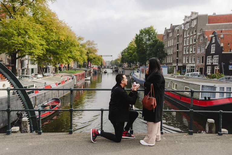 Man proposes on an Amsterdam canal bridge as his partner reacts with joy. Boats and historic houses frame the moment during a surprise engagement session in the city.