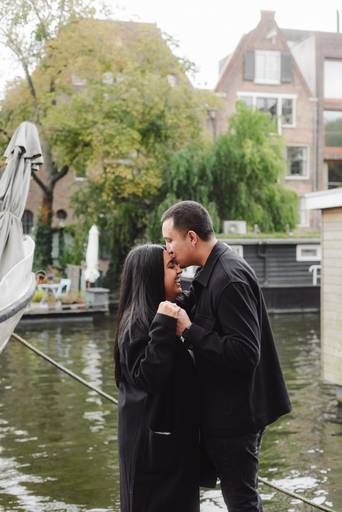 Couple embraces by the water in Amsterdam after their surprise engagement, standing near houseboats and historic buildings.