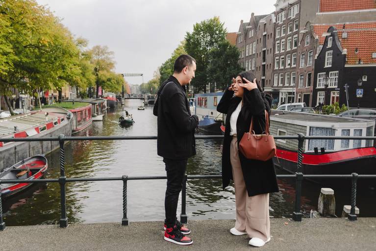 Couple stands on an Amsterdam bridge, smiling and talking right after their surprise engagement, with boats and canal houses behind them.