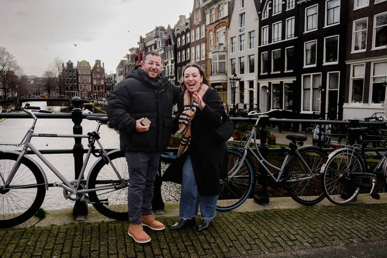 Couple on a canal bridge reacting joyfully after the proposal, with Amsterdam houses and bicycles behind them.