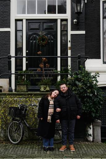 Couple standing in front of a black door with a winter wreath and a bicycle nearby, the woman leaning her head on the man’s shoulder.