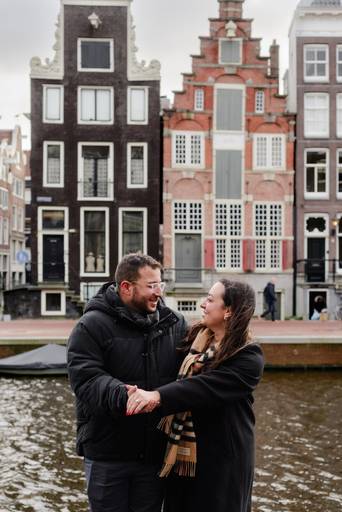 Couple holding hands and smiling at each other on a canal bridge with traditional Amsterdam houses behind them.