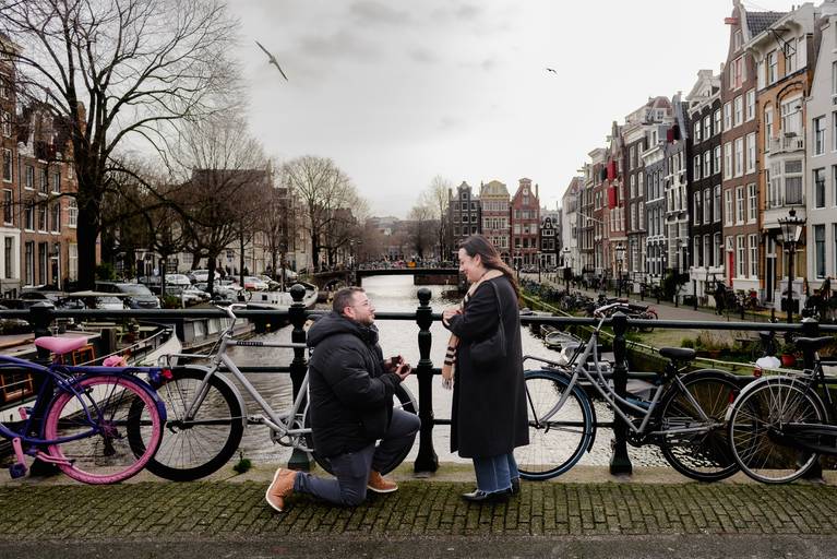 Man kneeling on a canal bridge proposing, while the woman smiles and looks at him with Amsterdam’s canal and houses in the background.