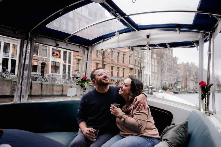 Couple sitting together on a boat with clear panels, holding drinks and smiling while looking up at the canal houses around them.