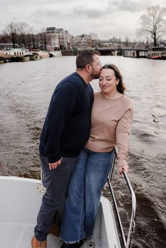 Couple standing at the front of a small boat on an Amsterdam canal, the man kissing the woman’s forehead while she smiles with her eyes closed.