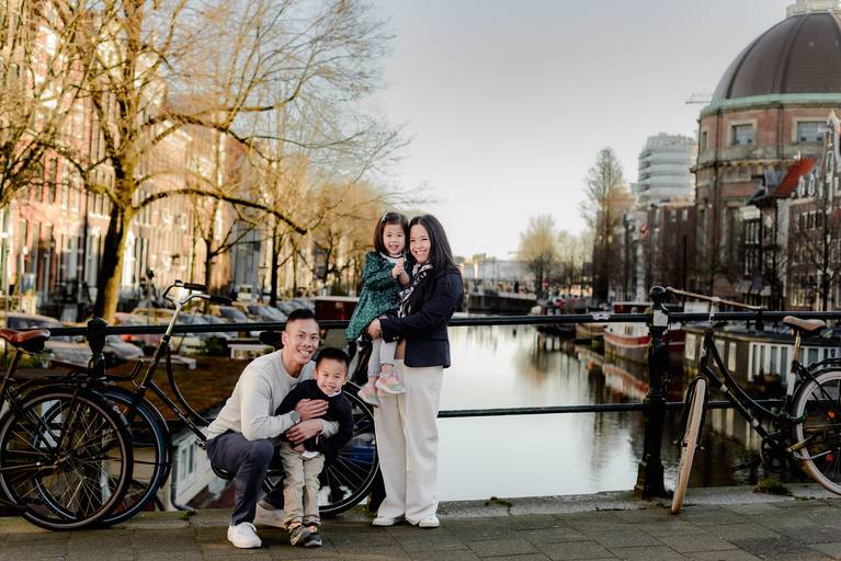 Family portrait on a canal bridge in Amsterdam, parents with two young children surrounded by bicycles and historic canal houses in soft morning light.