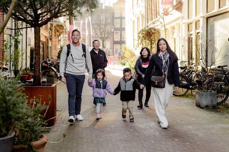 Extended family walking hand in hand through a quiet Amsterdam street, enjoying a relaxed vacation moment among historic buildings and greenery.