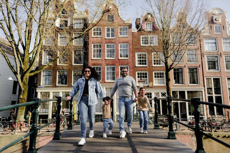 Family walking hand in hand across a small canal bridge in Amsterdam, captured during a joyful vacation photography session with traditional Dutch houses behind them.