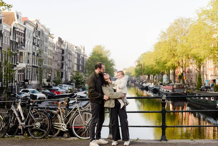 Family with a baby standing on a bridge along an Amsterdam canal, surrounded by bicycles and historic canal houses in spring.