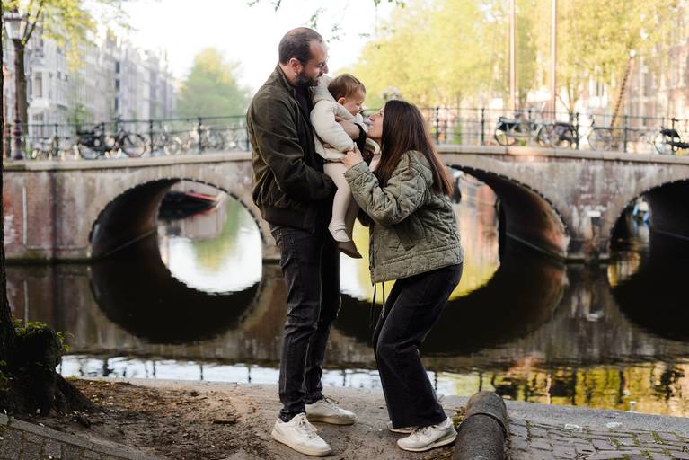 Parents holding their baby near a small stone bridge over an Amsterdam canal, sharing a quiet family moment outdoors.