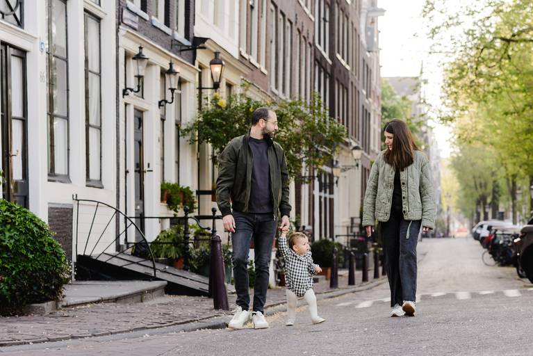 Parents walking hand in hand with their toddler on a quiet residential street in Amsterdam, with canal houses and greenery in the background.