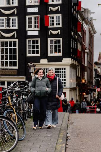 Two women walking arm in arm in Amsterdam, smiling near parked bicycles and historic canal houses in winter.
