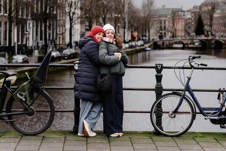 Mother and daughter hugging on an Amsterdam canal bridge, bicycles on both sides and classic canal houses in the background.