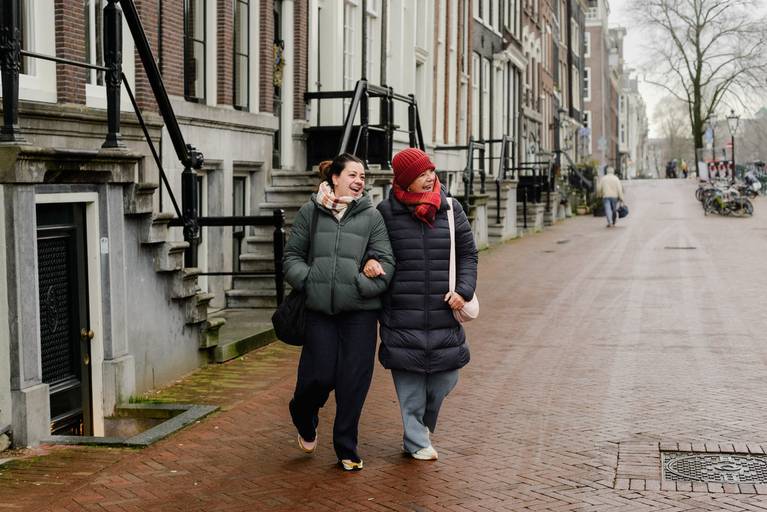 Mother and daughter laughing while walking together on a brick street in Amsterdam, surrounded by traditional canal houses.