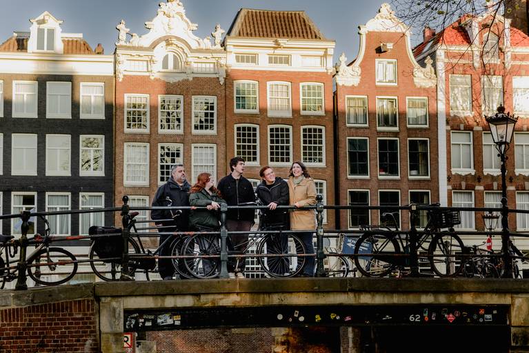 Family group leaning on a canal bridge railing in Amsterdam, framed by classic Dutch canal houses and bicycles, enjoying a relaxed moment during a family photo session.