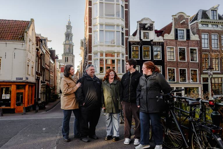 Multi-generation family standing together on a bridge in Amsterdam, smiling and talking, with the Westertoren visible in the background and historic canal houses around them.