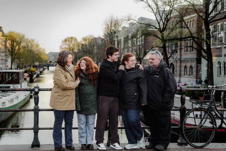 Extended family posing closely together on an Amsterdam canal bridge at sunset, with boats on the water and tree-lined canals creating a calm, everyday city atmosphere.
