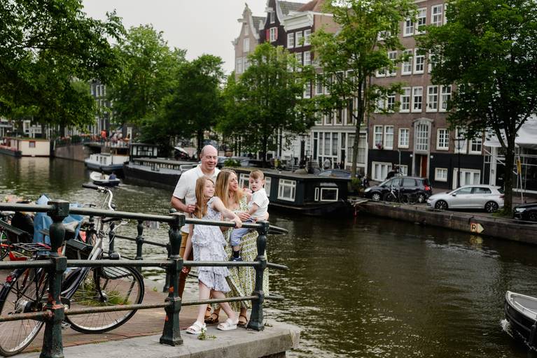 Family standing together by an Amsterdam canal, surrounded by houseboats, trees, and traditional canal houses on a summer day.