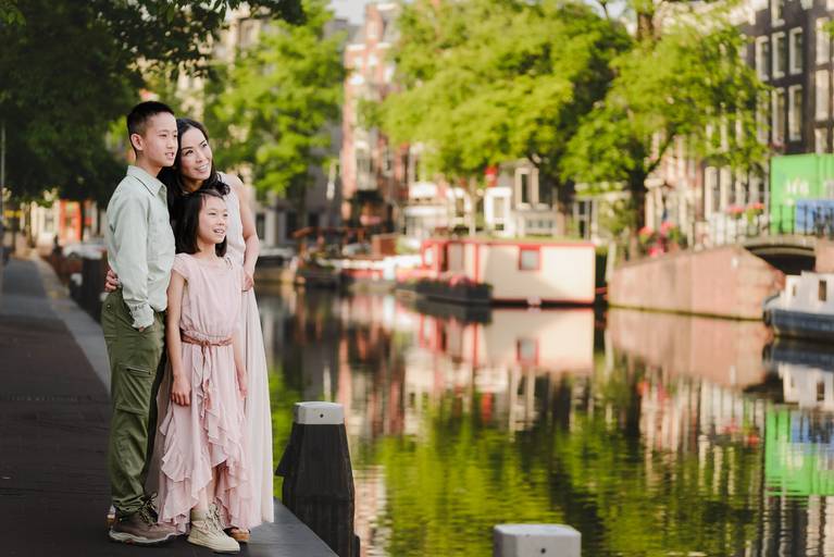 Mother with two children standing by an Amsterdam canal, looking out over the water with houseboats and trees in the background.