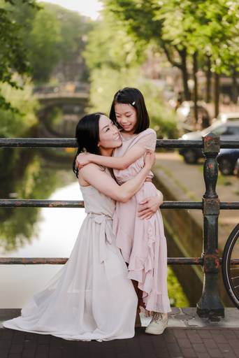 Mother hugging her daughter on a bridge over an Amsterdam canal, captured in soft evening light during a family photo session.
