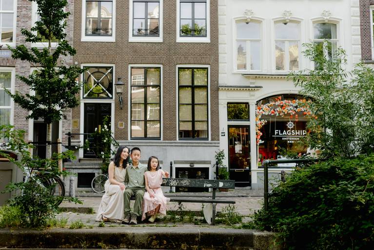 Mother and two children sitting on a bench by an Amsterdam canal with historic canal houses and greenery in the background.