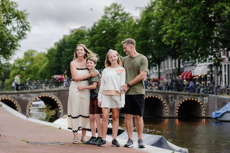 Family of four standing together by an Amsterdam canal with a stone bridge in the background during an outdoor photo session.