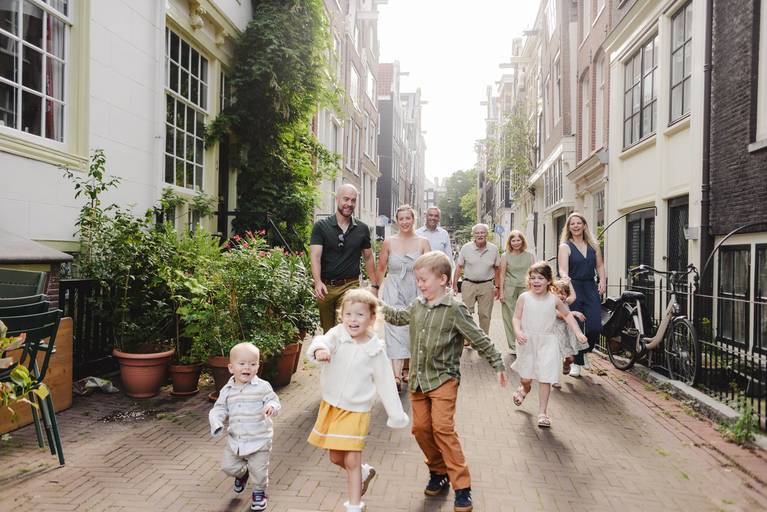 Large multi-generation family walking together down a quiet Amsterdam street as young children run ahead, surrounded by canal houses and greenery.