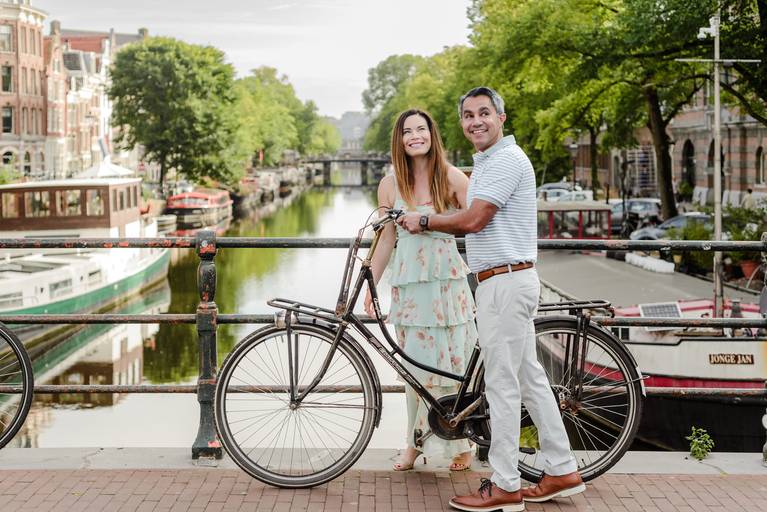 Couple standing with a Dutch bike on an Amsterdam bridge, enjoying canal views during their anniversary trip to the city.