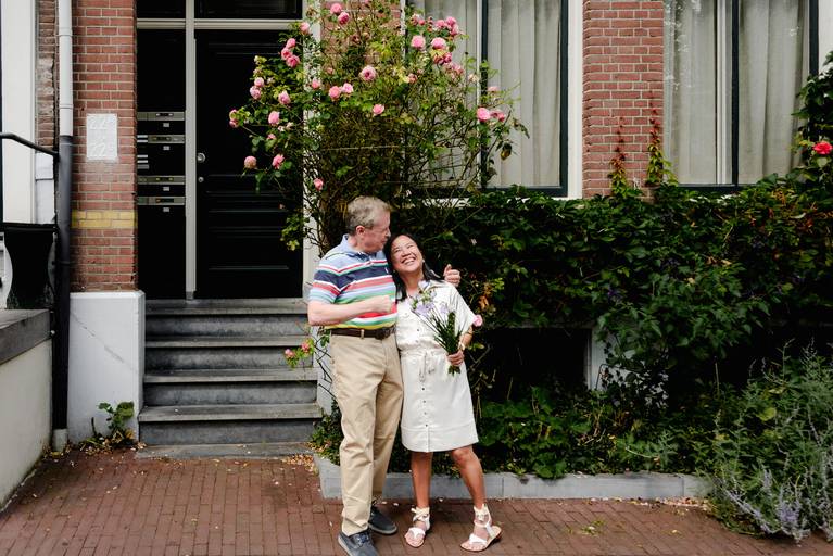 Couple laughing together in front of an Amsterdam home covered in summer flowers during their travel photoshoot.