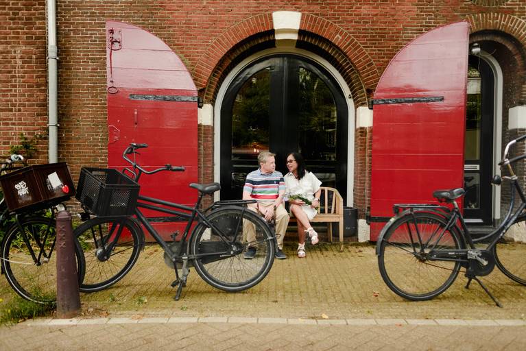 Couple sitting outside a historic Amsterdam building with red shutters, enjoying a relaxed vacation photo session.