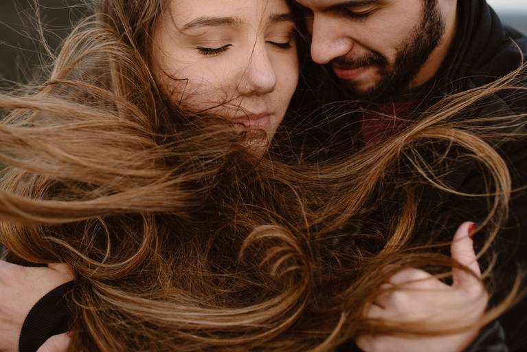 Ensaio pré wedding, os noivos estão abraçados, totalmente envolvidos e o vento faz o cabelo da noiva se movimentar e envolvê-los.
