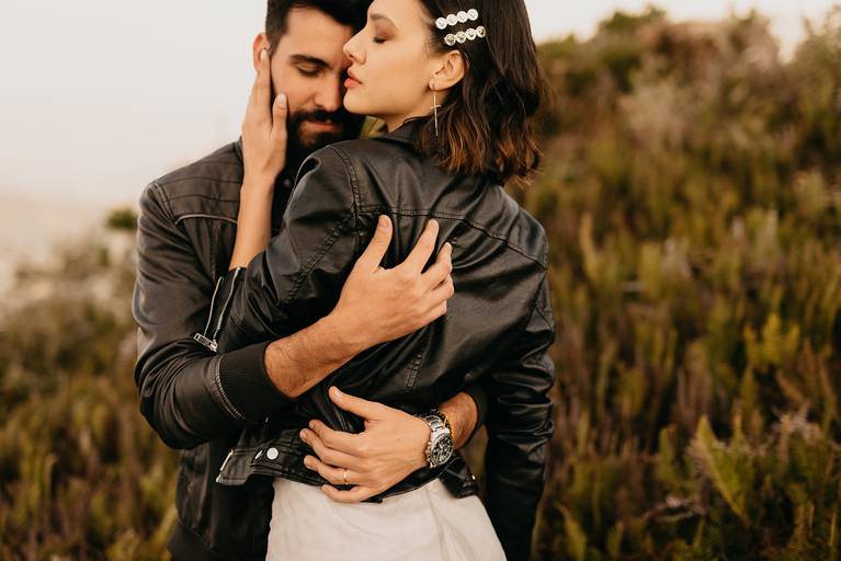 Noivos de jaqueta de couro preta no alto da montanha, o noivo abraça a noiva com muita paixão pelas costas e a noiva toca o noivo no rosto com muita delicadeza. 