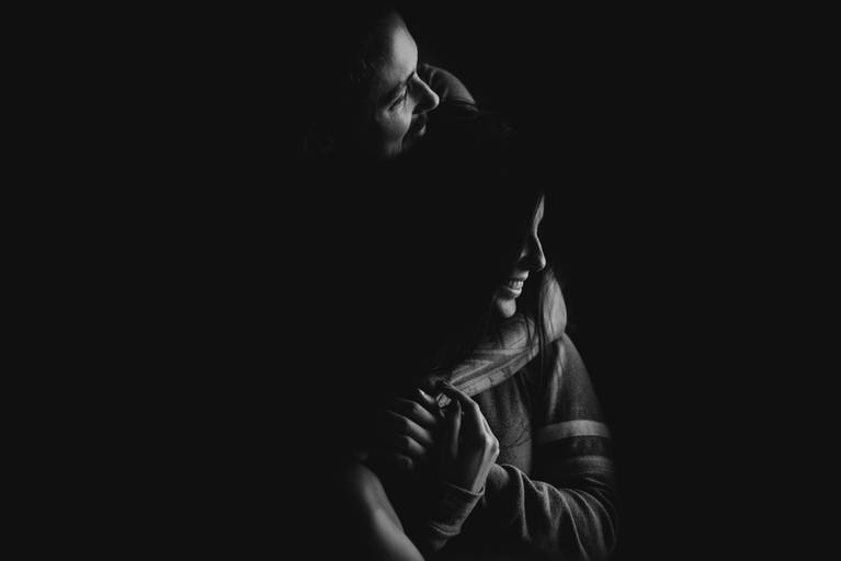 foto preto e branca, com luz de janela iluminando somente o rosto do casal que está abraçado e conectado. 