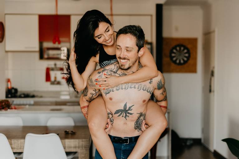 noiva sorrindo e muito feliz nas costas do seu noivo, noivo tatuado e sem camisa no ensaio pré-casamento em casa.