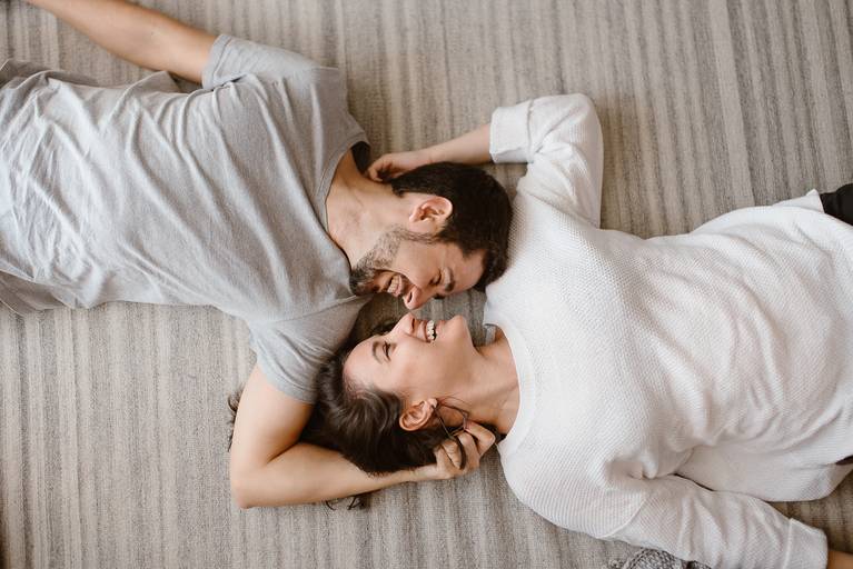 Os noivos estão deitados no chão, sorrindo, e muito apaixonados em um ensaio em casa.