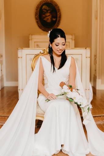 retrato de uma noiva oriental, ela está sentada em uma cadeira de madeira com detalhes em dourado e arabescos, ela está segurando um buque de orquídeas no colo e sorrindo. 