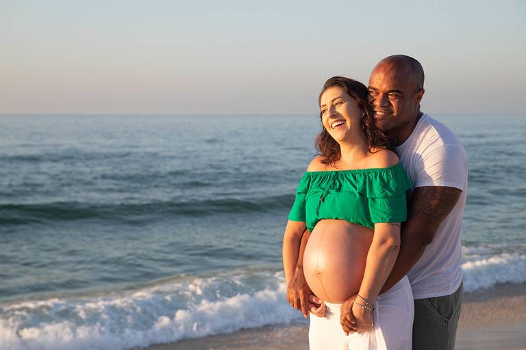 Casal de gestante abraçado à beira-mar em ensaio na Praia da Barra.