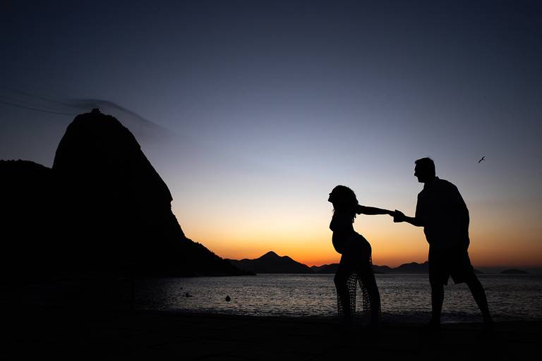 Casal de gestantes na Praia Vermelha, com sol se pondo atrás do Pão de Açúcar.