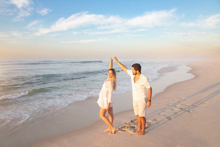 Casal de gestantes caminhando na areia com o mar batendo nos pés.