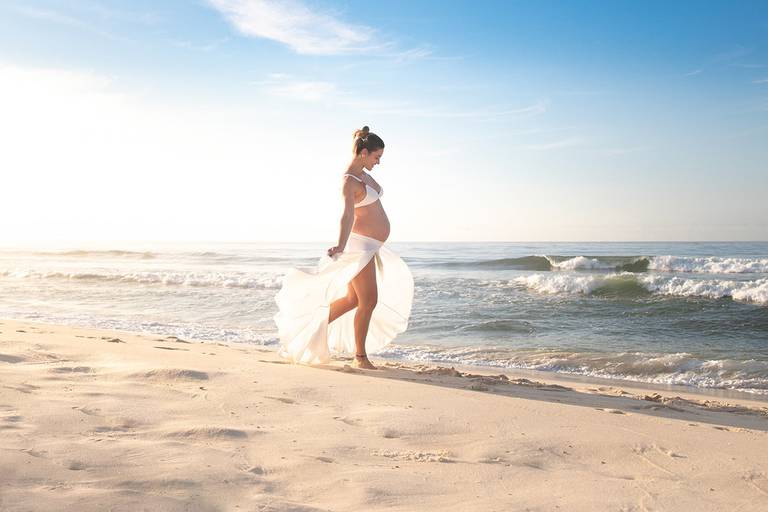 Gestante caminhando sozinha na praia da Barra com vestido branco.